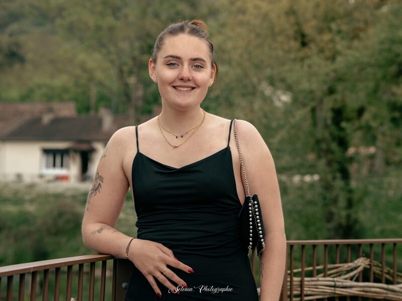 Portrait d’une jeune femme en robe noire, posant et souriant sur une terrasse avec un décor verdoyant en arrière-plan.