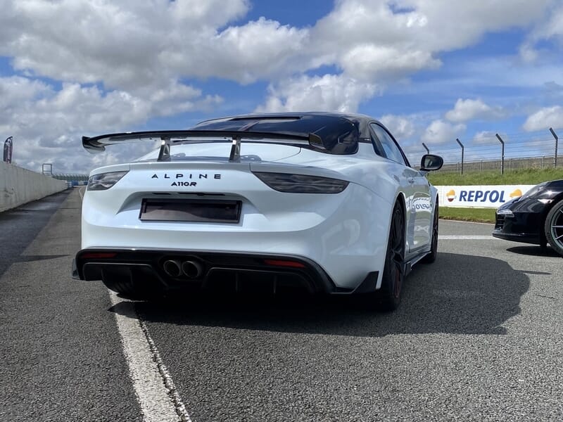 Alpine A110R blanche et noire de dos sur piste, aileron carbone et échappement central.