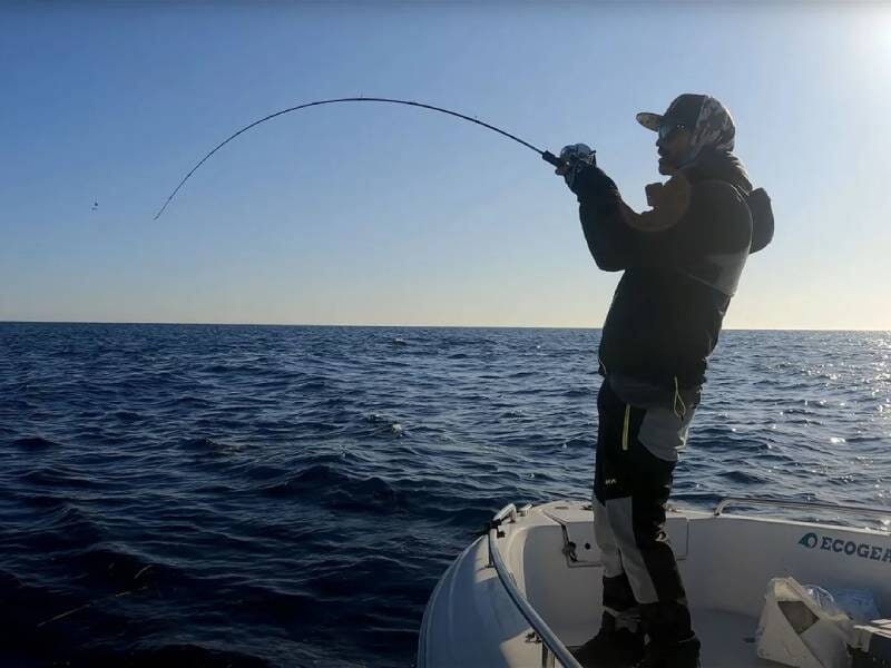 Un homme debout sur un bateau tient une canne à pêche arrondie par le poids d'un poisson qu'il est en train de remonter.
