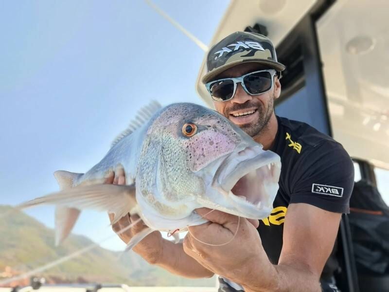 Un homme avec une casquette et des lunettes de soleil pose avec un poisson blanc et irisé dans les mains.