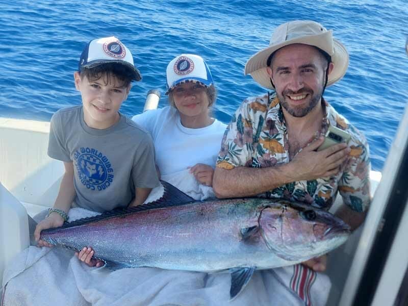 Deux enfants et un homme souriant posent sur un bateau avec un grand poisson pêché en mer.