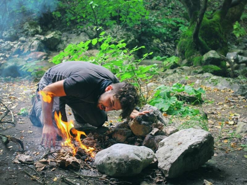 Un homme accroupi allume un feu de camp avec des brindilles et des pierres dans un environnement forestier, près de Vannes.