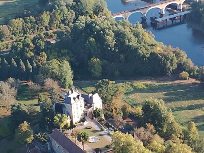 Château en pierre entouré de verdure avec un pont enjambant la rivière Dordogne.