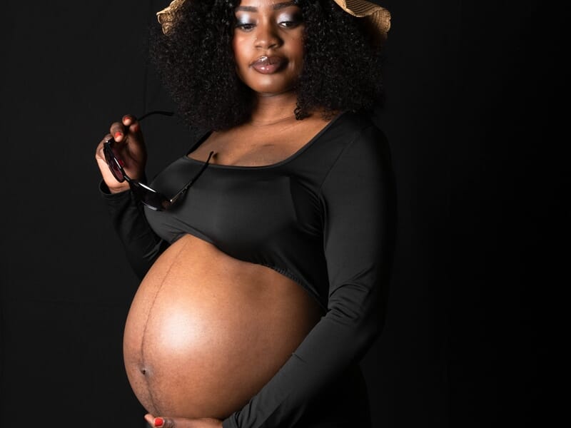 Future maman en robe noire, accessoirisée d’un chapeau, regard affirmé.