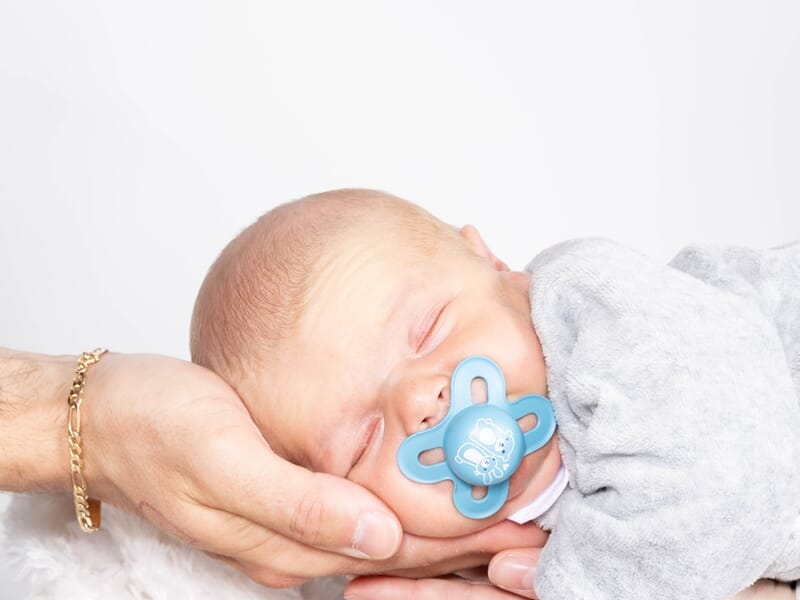 Portrait tendre d’un bébé endormi, appuyé dans les mains de ses parents, tétine bleue à la bouche.