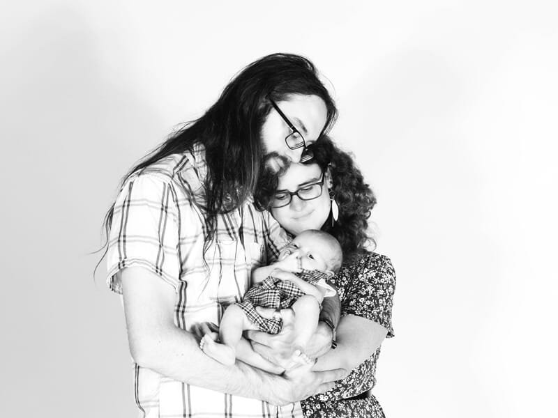 Jeune famille posant en studio, parents enlacés tenant leur nourrisson avec douceur en noir et blanc.