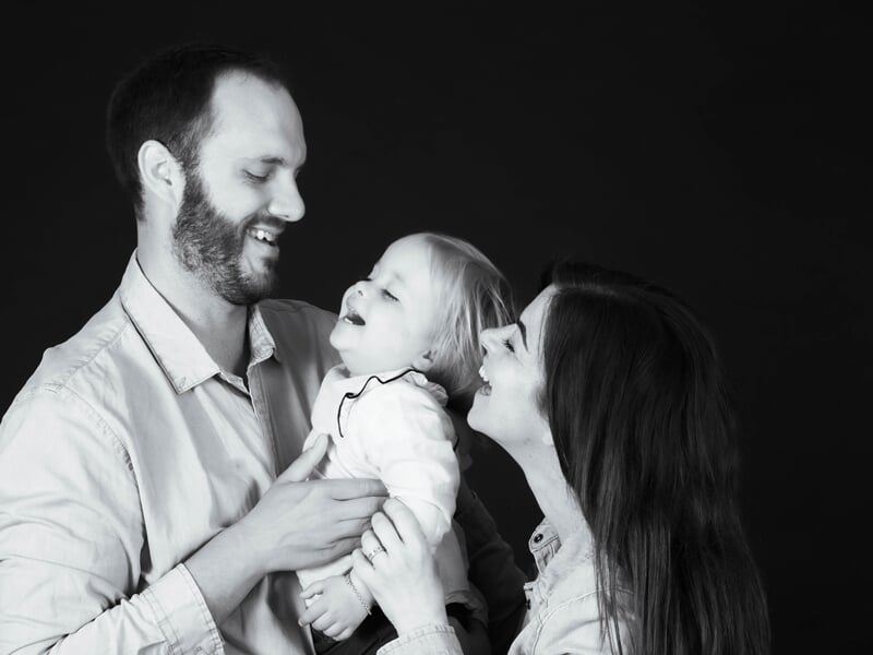 Jeune famille en studio, parents souriants jouant avec leur enfant dans une ambiance tendre en noir et blanc.