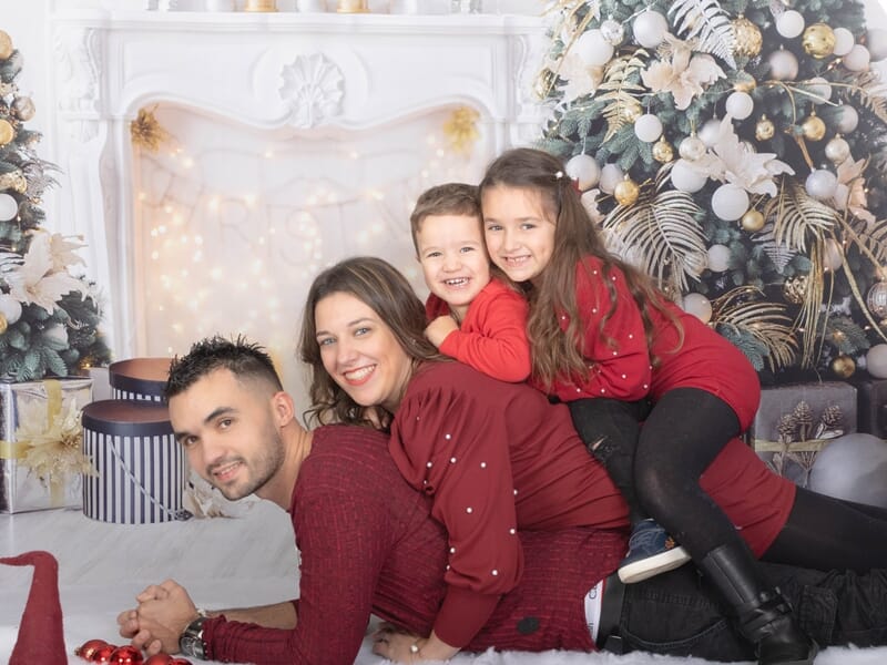 Famille en tenue rouge posant joyeusement allongée devant une cheminée décorée de Noël avec sapin et guirlandes lumineuses.
