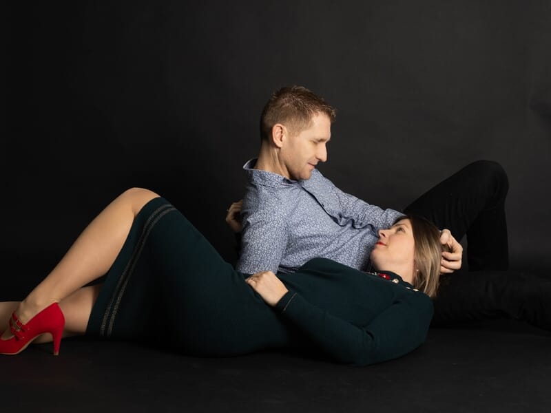 Couple allongé au sol dans un studio, l’homme en chemise bleue regarde tendrement la femme en robe verte et talons rouges qui repose sa tête sur lui.