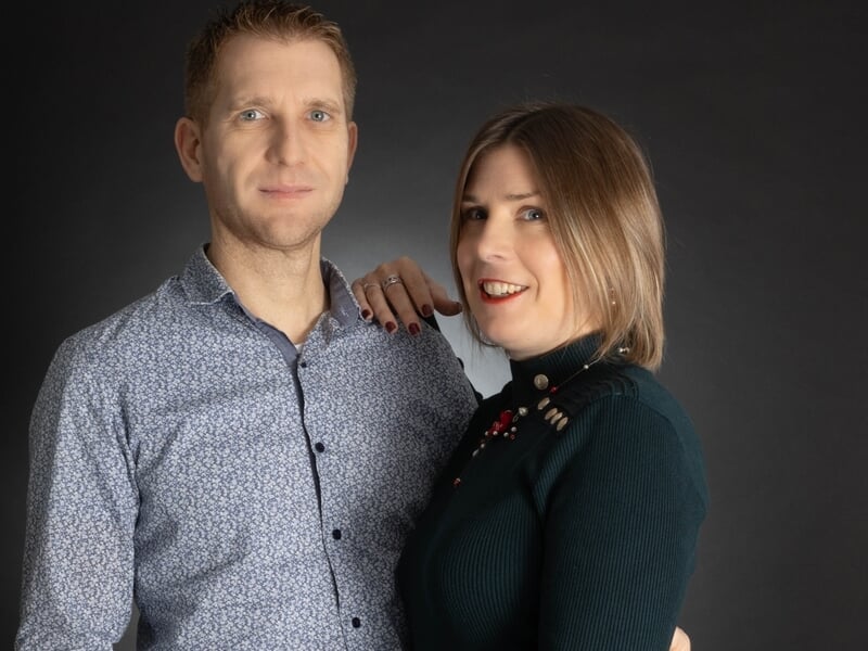 Couple posant ensemble en studio, l’homme en chemise bleue et la femme en robe verte, se tenant affectueusement par la main devant un fond noir.