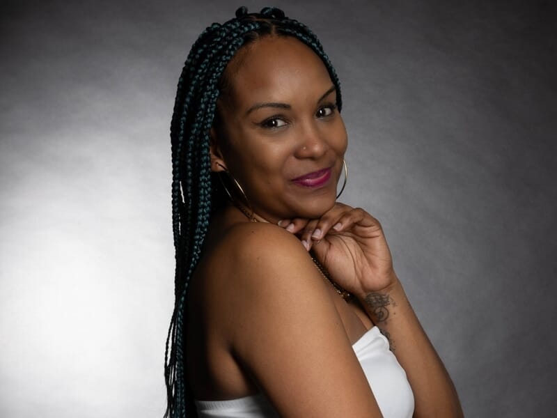 Femme souriante aux tresses, posant en studio avec un haut blanc et un fond sombre.
