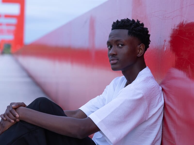 Portrait d’un jeune homme assis contre un mur rouge, vêtu d’une chemise blanche, ambiance urbaine.