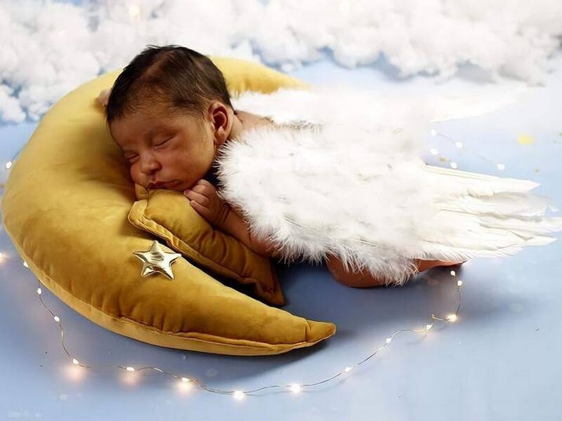 Bébé endormi sur un coussin en forme de lune dorée, avec des ailes d’ange blanches et guirlande lumineuse autour, image prise par une photographe bébé à Dijon.