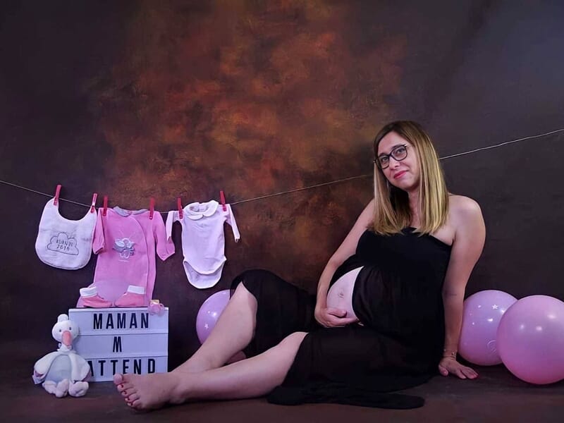 Femme enceinte assise en robe noire transparente, entourée de vêtements et accessoires roses pour bébé, image prise lors d'un shooting photo grossesse à Dijon