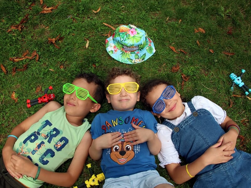 Séance photo famille Dijon en plein air, trois enfants allongés sur l’herbe avec lunettes colorées, éclats de rires et ambiance estivale.