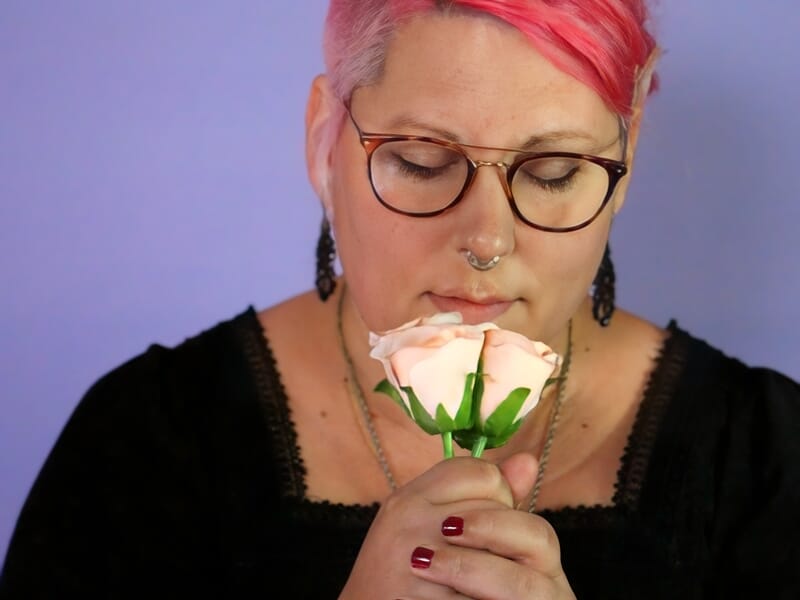 Séance photo portrait Dijon, femme aux cheveux roses et lunettes tenant une rose avec douceur sur fond violet.