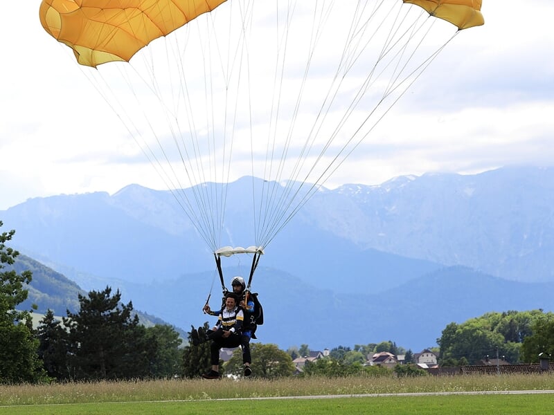Personne en parachute jaune atterrissant sur un champ vert avec montagnes et ciel nuageux en arrière-plan