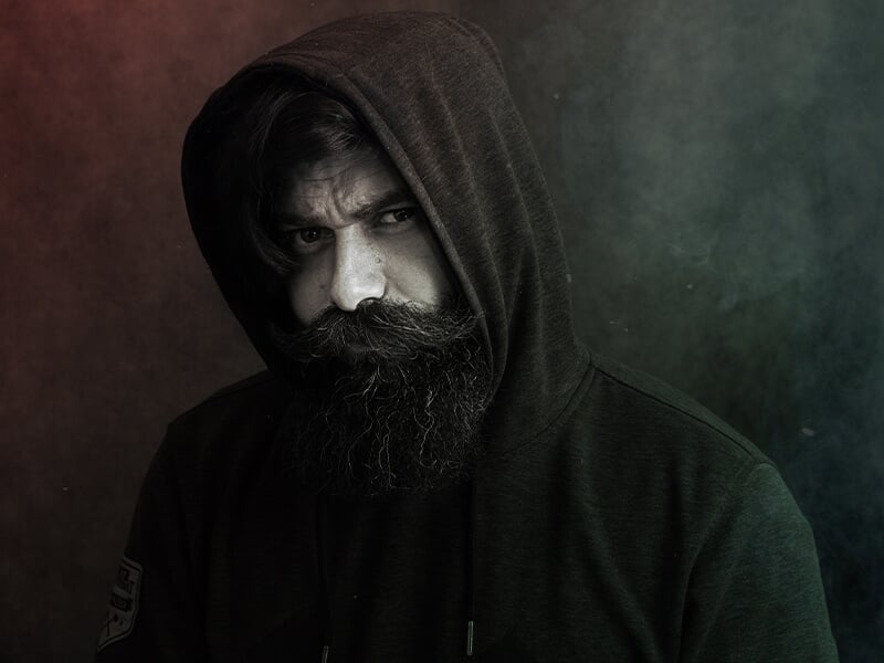 Photographe portrait Angers, homme barbu en sweat à capuche sombre, regard intense dans une ambiance artistique.