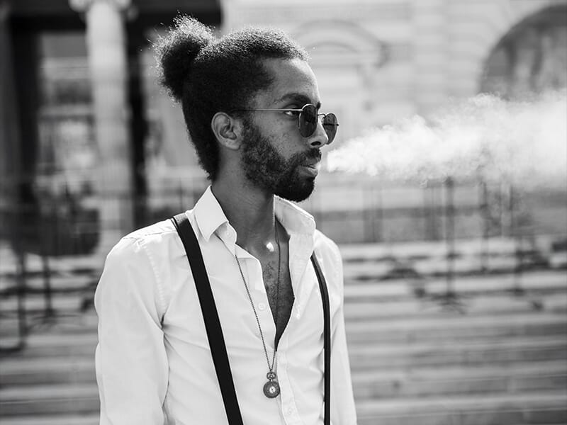 Photographe portrait Angers, homme élégant avec lunettes et bretelles, exhalant de la fumée dans un style urbain noir et blanc.