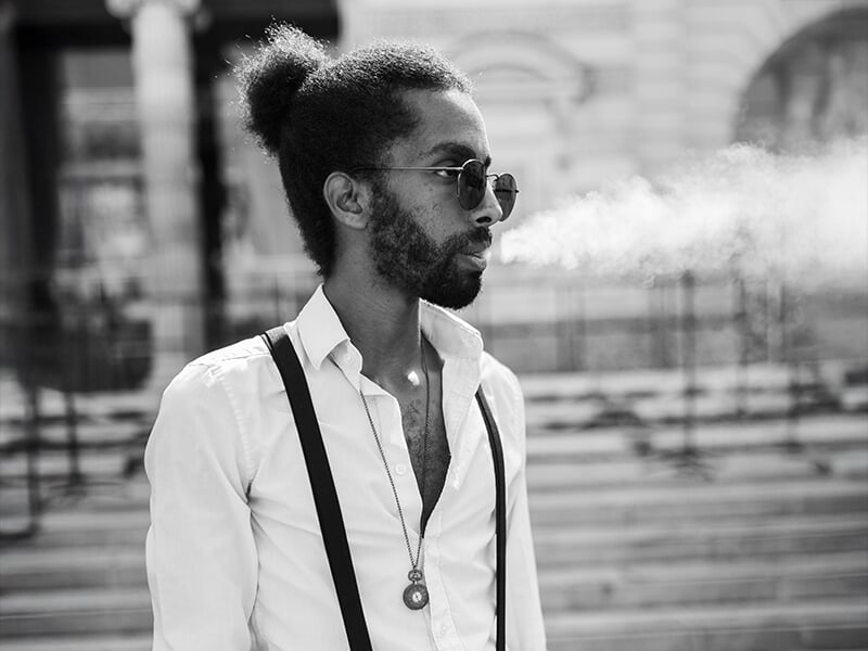 Photographe portrait Angers, homme élégant avec lunettes et bretelles, exhalant de la fumée dans un style urbain noir et blanc.