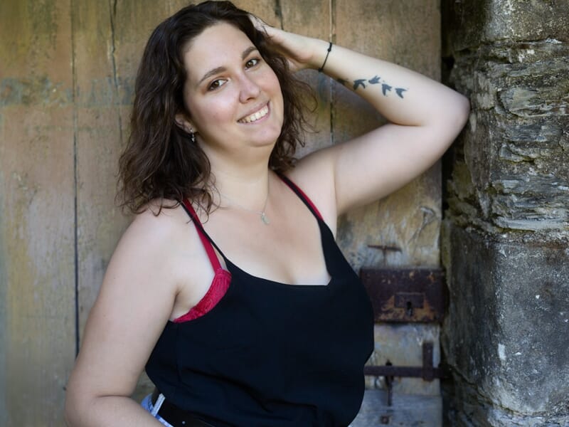 Photographe portrait Angers, jeune femme souriante posant contre un vieux mur en pierre, tatouage d’oiseaux visible sur le bras.