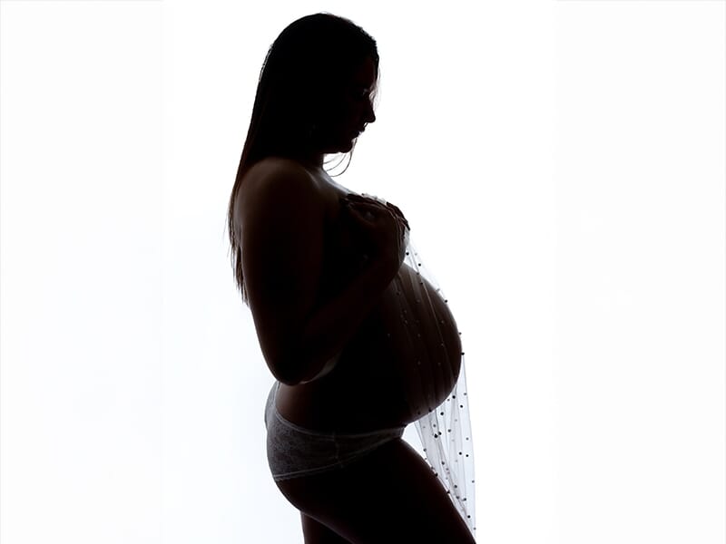 Silhouette en contre-jour d'une femme enceinte tenant son ventre, avec un tissu transparent orné de perles lors d'une séance photo grossesse à Angers.