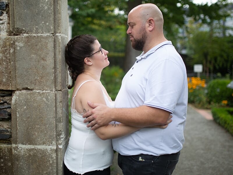 Un couple se regarde tendrement, l'homme en polo blanc et la femme en débardeur blanc, appuyés contre un mur en pierre lors d'un shooting avec une photographe couple à Angers.