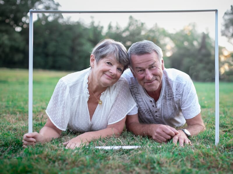 Un couple âgé souriant est allongé dans l'herbe verte, encadré par un cadre blanc lors d'un shooting avec une photographe couple à Angers.
