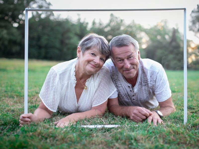Un couple âgé souriant est allongé dans l'herbe verte, encadré par un cadre blanc lors d'un shooting avec une photographe couple à Angers.