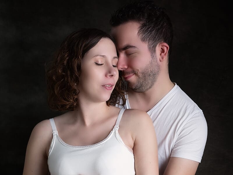 Un couple jeune pose en studio sur fond noir, la femme aux yeux fermés et l'homme souriant lors d'un shooting avec une photographe couple à Angers.