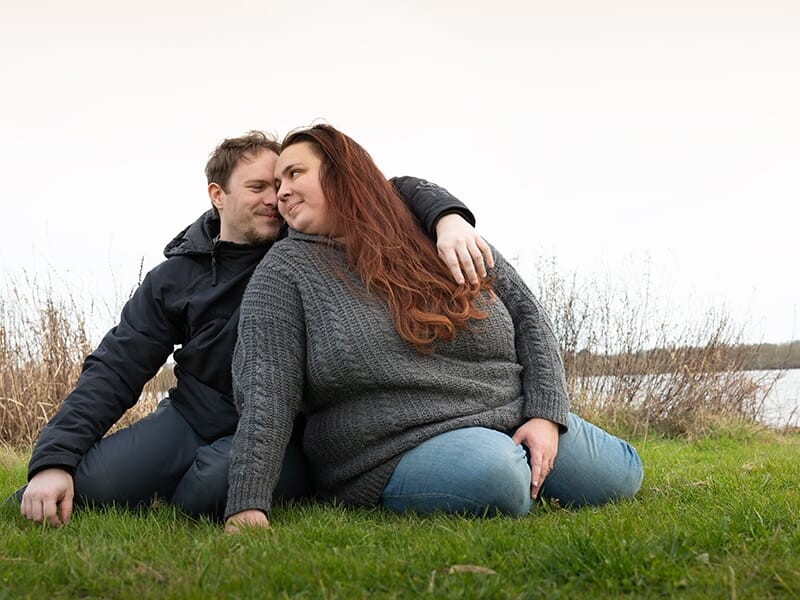 Un couple assis sur l'herbe près d'un plan d'eau, l'homme enlace la femme aux longs cheveux roux lors d'un shooting avec une photographe couple à Angers.