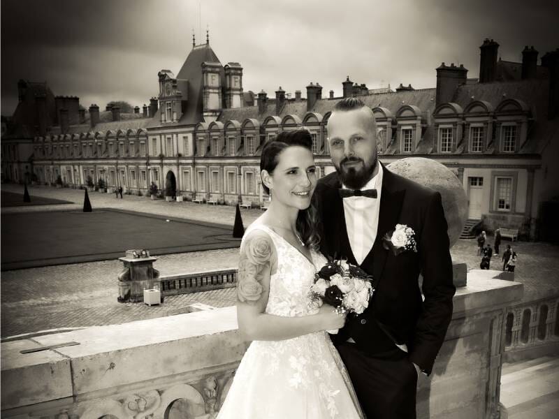 Photo en noir et blanc d'un couple de mariés posant devant un grand bâtiment historique avec un ciel nuageux, près d'Argelès.