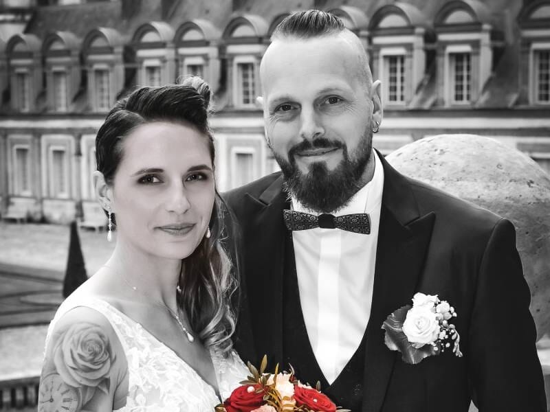 Couple de mariés en noir et blanc, la femme avec un tatouage sur le bras et le marié portant un nœud papillon et une boutonnière.