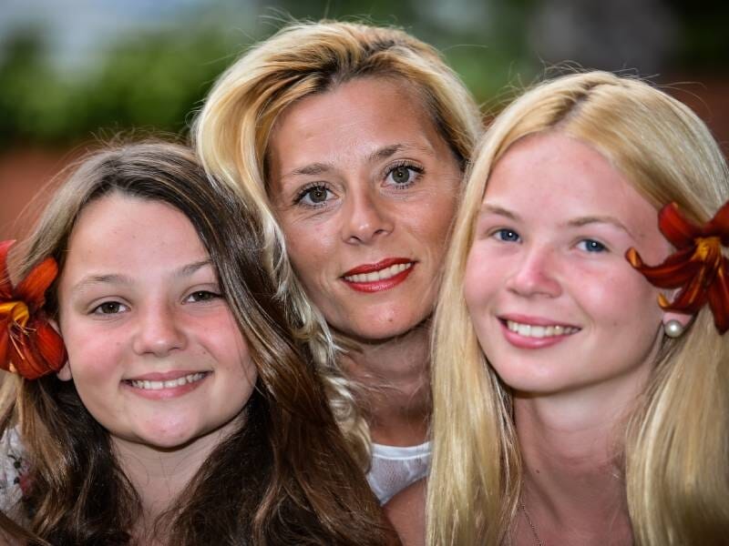 Gros plan de trois femmes souriantes, deux avec des fleurs rouges dans les cheveux, en arrière-plan flou.