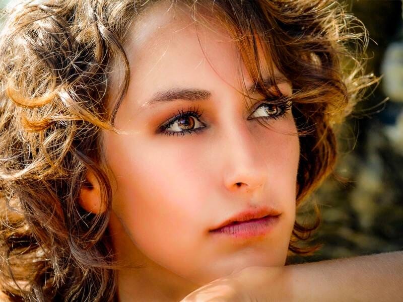 Jeune femme aux cheveux bouclés et regard pensif, éclairage naturel mettant en valeur son visage.