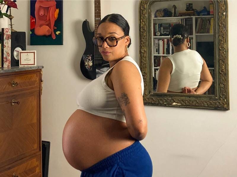 Une femme enceinte prend la pose, vêtue d'une brassière blanche et d'un pantalon bleu roi.