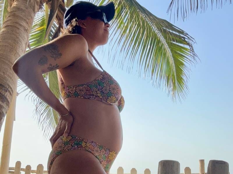 Une femme enceinte en maillot de bain regarde au loin, les mains sur les hanches. Elle pose devant un palmier.