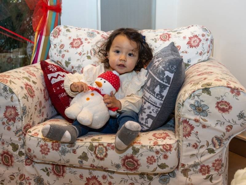 Une petite fille prend la pose avec une peluche bonhomme de neige dans un grand fauteuil fleuri.