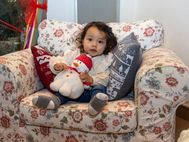 Une petite fille prend la pose avec une peluche bonhomme de neige dans un grand fauteuil fleuri.