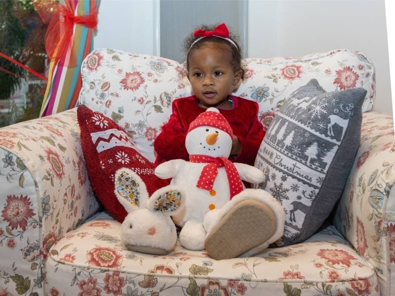 Une petite fille prend la pose avec une peluche bonhomme de neige dans un grand fauteuil fleuri.