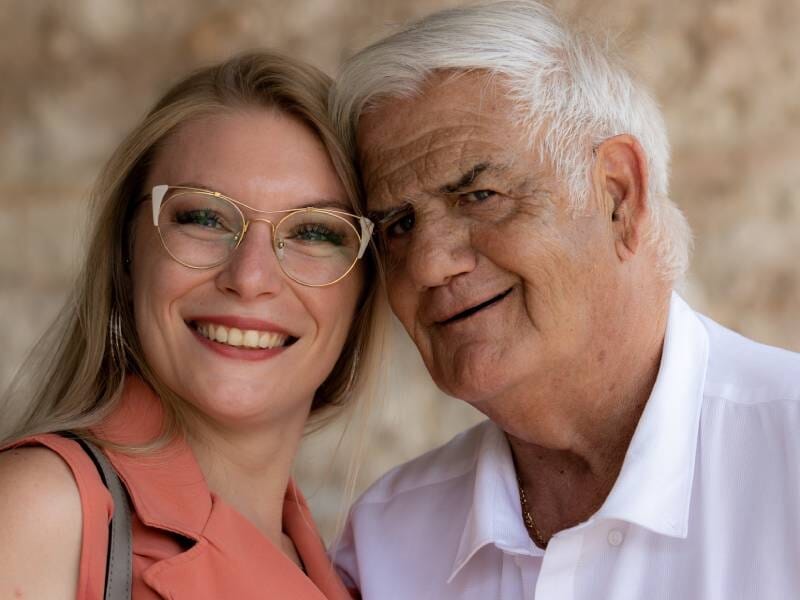 Portrait d'une femme souriante avec lunettes et d'un homme âgé souriant, posant ensemble devant un mur flou.