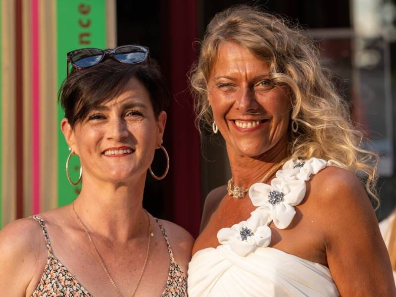 Portrait de deux femmes souriantes en extérieur, l'une porte une robe blanche avec fleurs, l'autre un top fleuri.