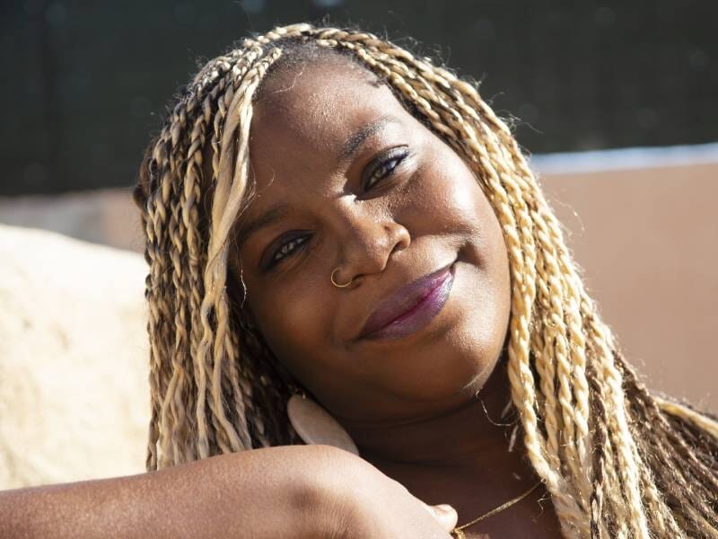 Portrait d'une femme souriante aux cheveux tressés blonds, avec un anneau au nez, posant en extérieur au soleil.