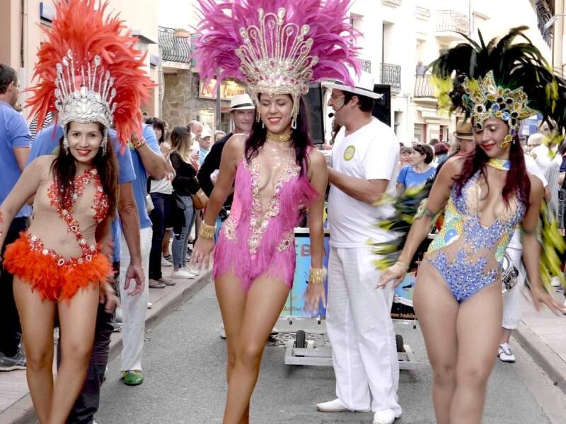 Trois danseuses en costumes colorés et plumes défilent dans une rue animée lors d'un carnaval ou festival.