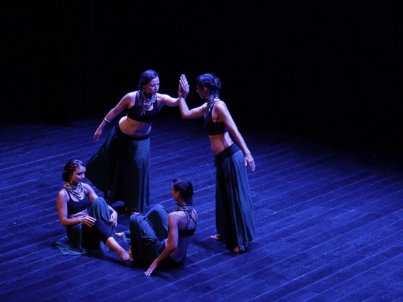 Quatre danseuses en tenue noire exécutent une chorégraphie contemporaine sur une scène sombre éclairée.