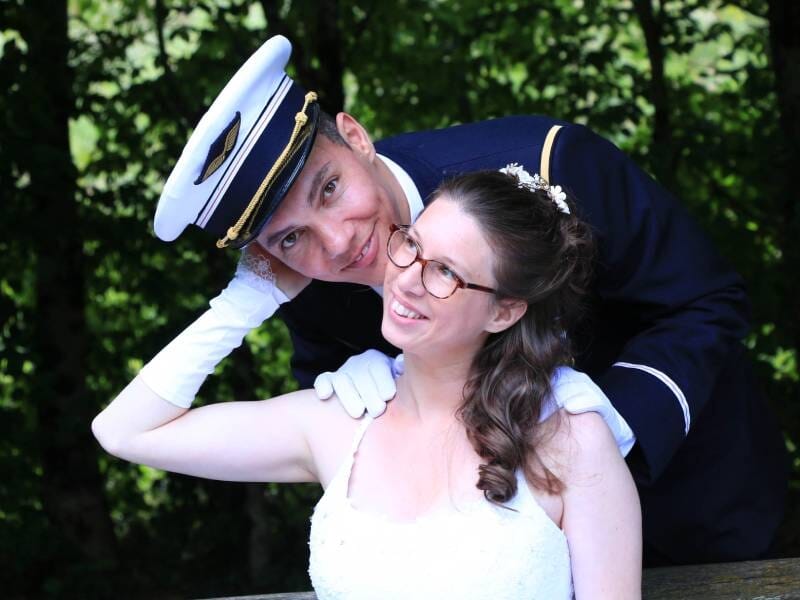 Un couple souriant en tenue de mariage, l'homme en uniforme militaire embrasse tendrement la femme en robe blanche.