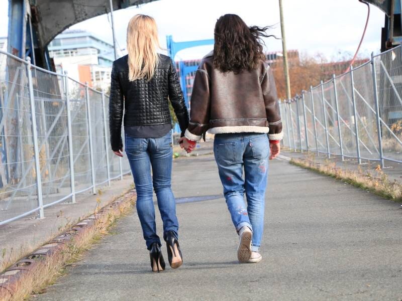 Deux femmes marchant main dans la main sur un chemin urbain, portant des vestes en cuir et des jeans décontractés.
