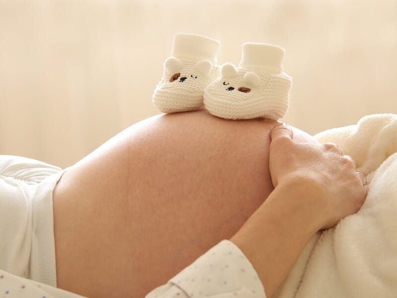 Ventre de femme enceinte avec chaussons de bébé blancs posés dessus, main caressant doucement le ventre arrondi.