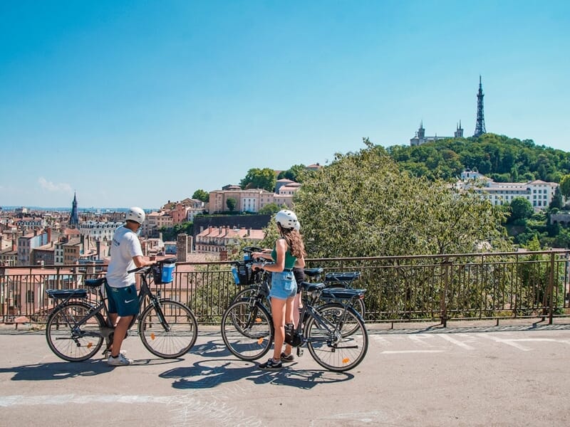Balade vélo électrique Lyon avec vue panoramique sur Fourvière, deux cyclistes en VAE profitent d’un arrêt face à la basilique et à la colline.