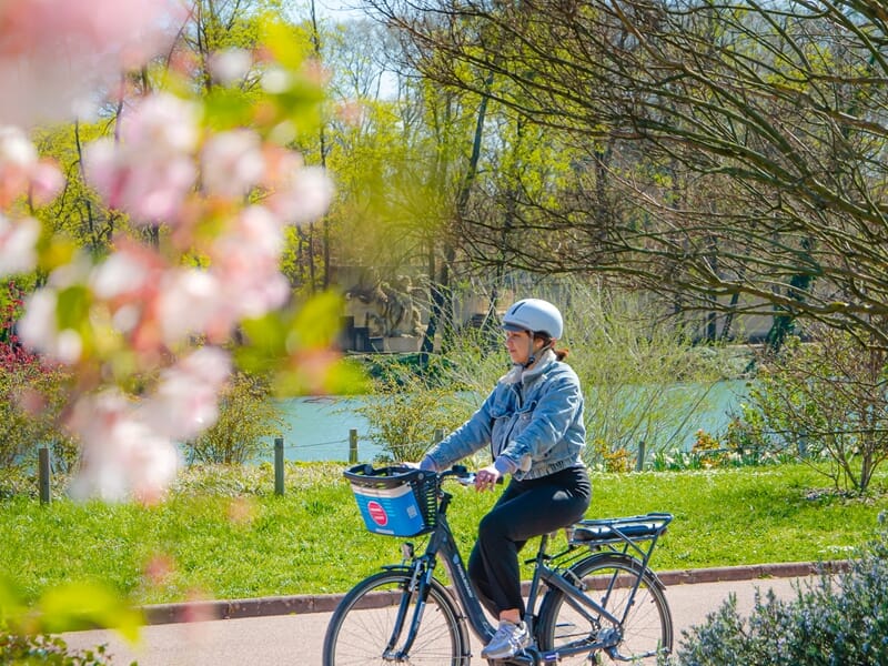 Femme en balade vélo électrique Lyon au parc, pédalant le long d’un chemin fleuri au bord de l’eau sous un ciel ensoleillé.